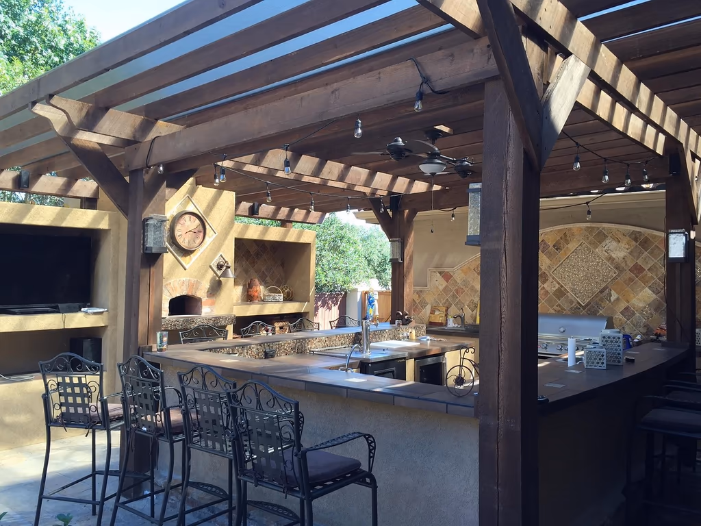 Covered outdoor kitchen with a timber pergola, wraparound bar seating, built-in grill, pizza oven, and TV for backyard entertaining.