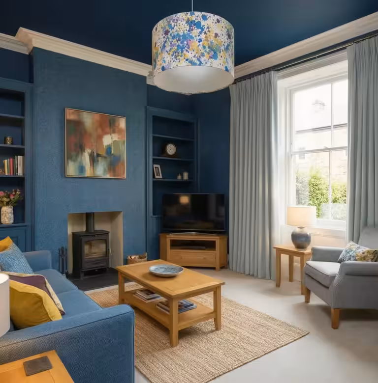 Contemporary living room with deep blue walls, cozy wood-burning stove, wooden furniture, large window with light curtains, colorful pendant light.