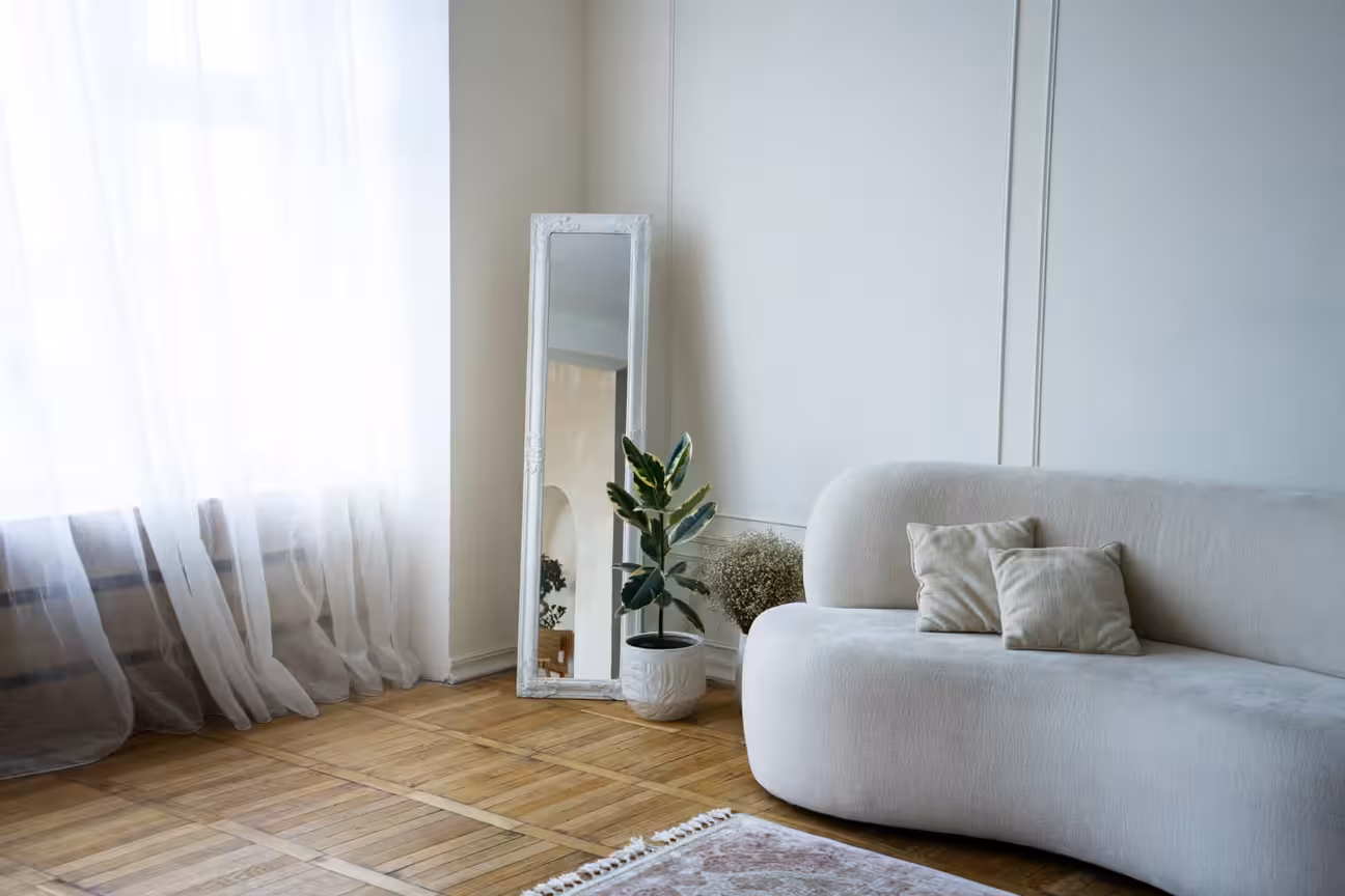 Minimalist living room with curved white sofa, floor mirror, indoor plant, and soft natural light through sheer curtains