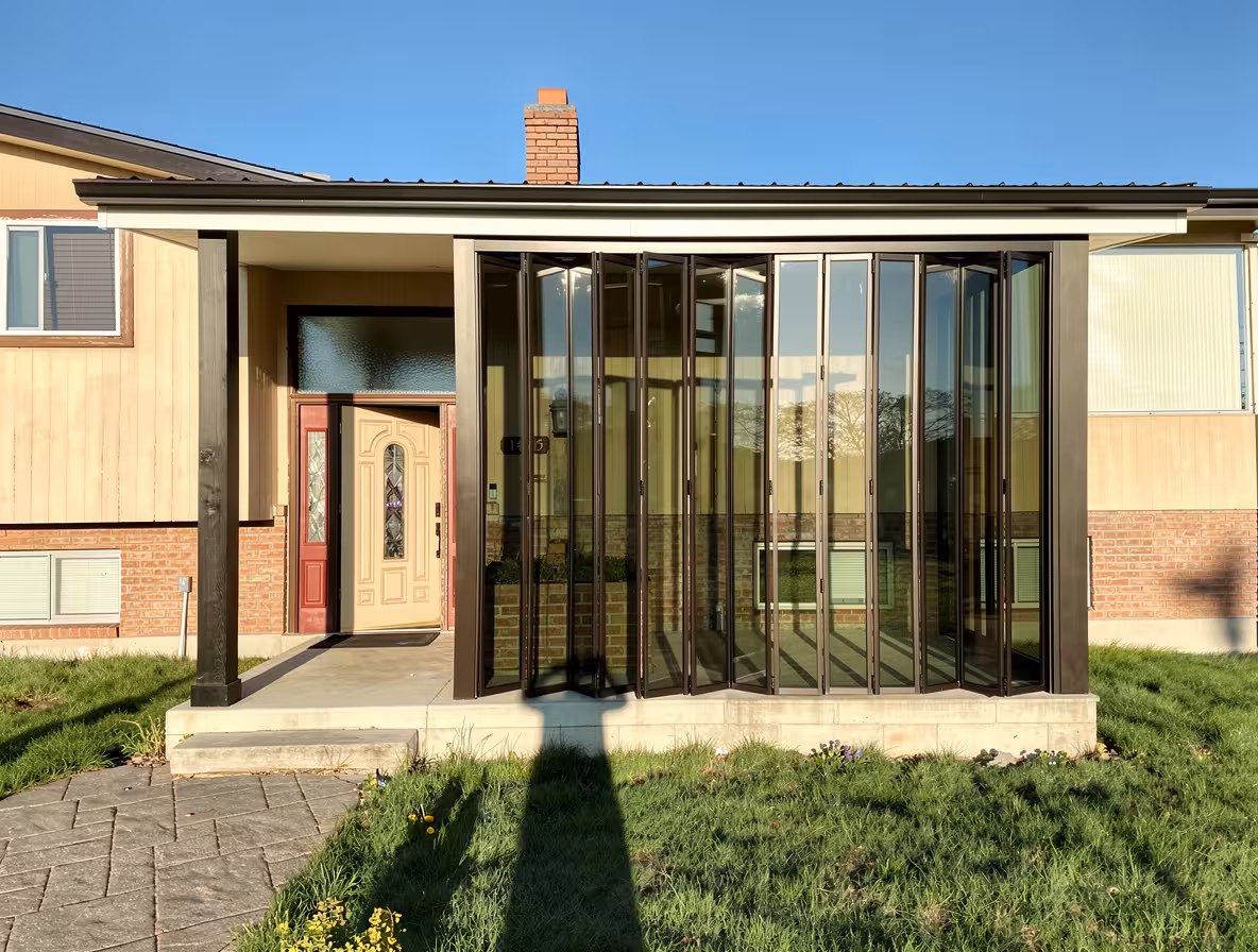 Contemporary enclosed porch with large folding glass panels, beige and red front door, brick and wood exterior, green lawn, and bright natural sunlight.
