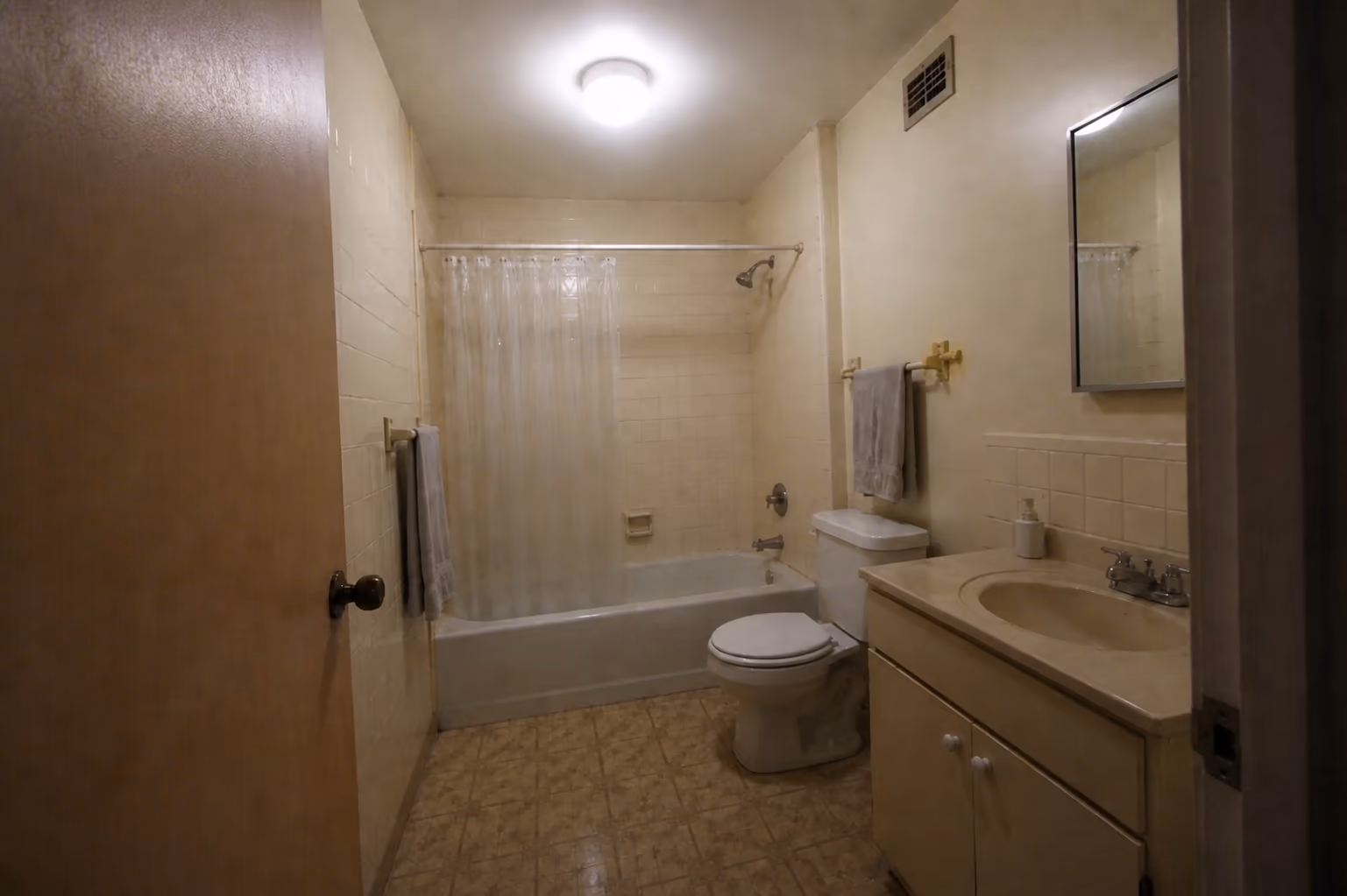 A lit photo of a dated bathroom with cream tile, a plastic shower curtain and a boxy vanity