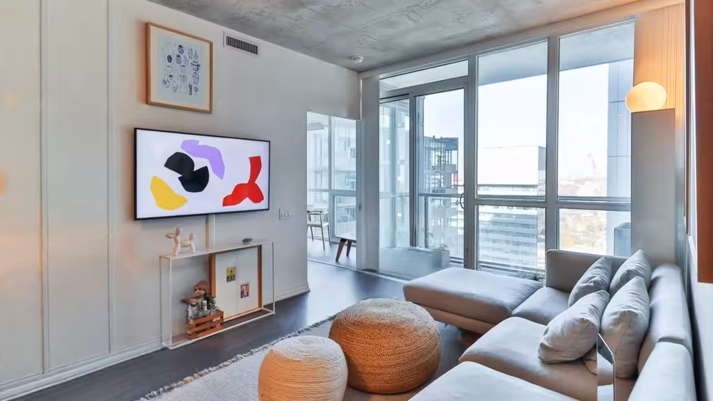 Contemporary living room with large windows, light gray sectional sofa, woven poufs, abstract wall art, concrete ceiling, and soft natural lighting.