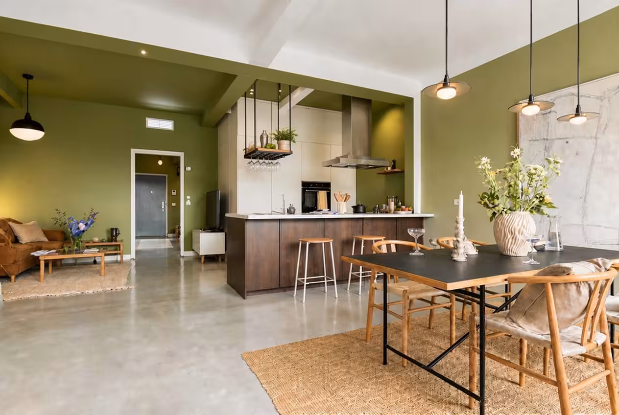 Modern open-plan dining and kitchen space with olive-green walls, wood furniture, and modern lighting.