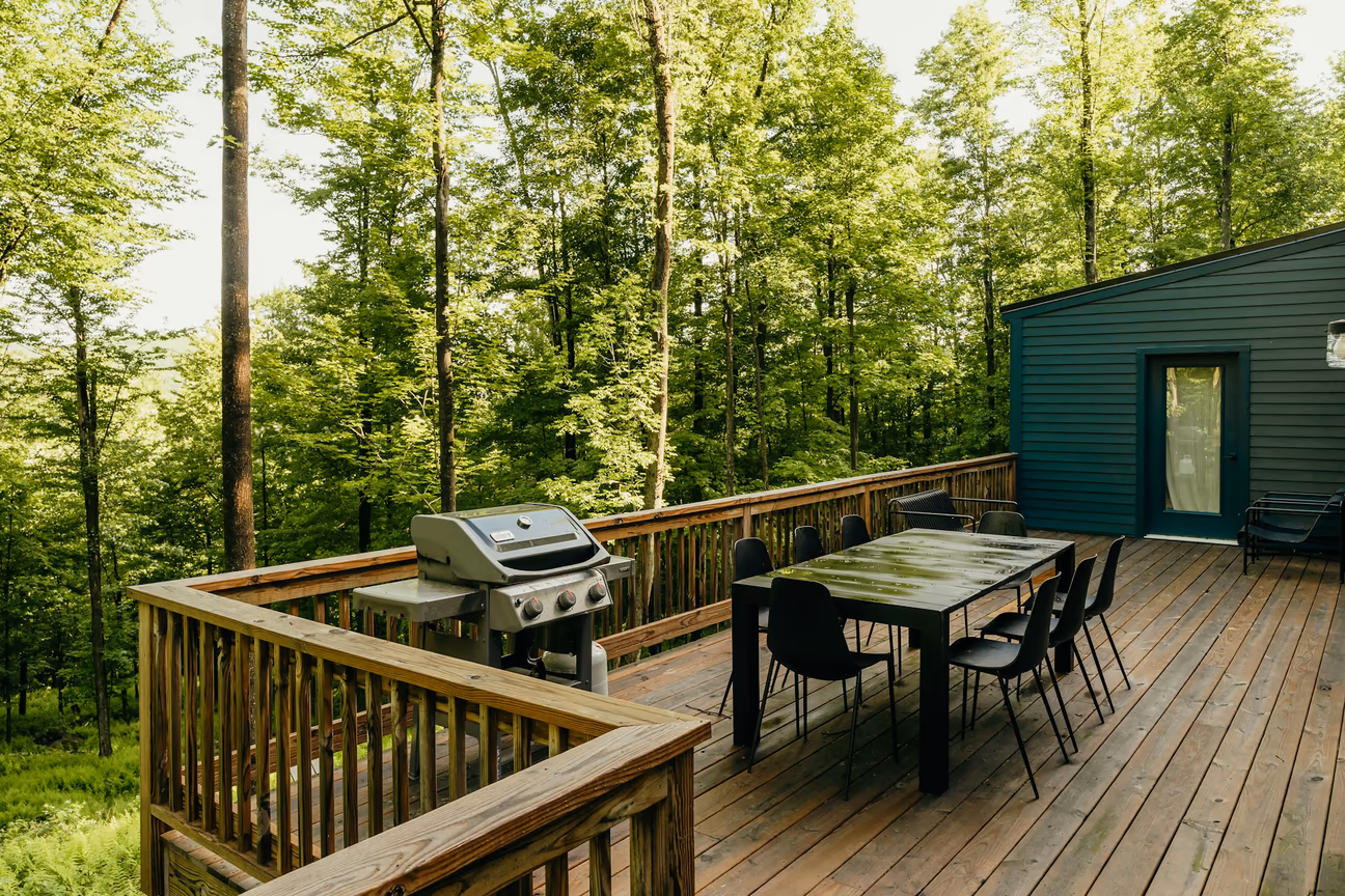 Rustic wooden deck outdoor kitchen with a gas grill, long dining table, and forest views surrounded by lush trees.
