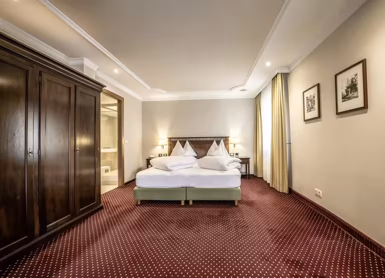 Traditional bedroom featuring a large wooden wardrobe, twin beds with white linens, beige walls, patterned red carpet, framed artwork, and soft warm lighting.