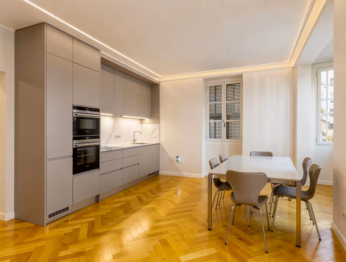 Modern kitchen with matte gray cabinetry, built-in stainless steel appliances, white marble backsplash, warm herringbone wood flooring, minimalist dining table, and soft ambient lighting.