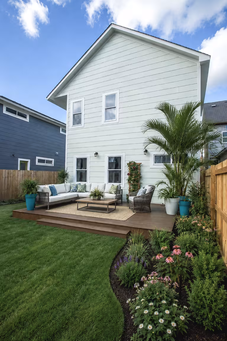 Raised dark wood deck with L-shaped sectional sofa, mint-green house, curved flower bed with coneflowers and salvia, and potted palms