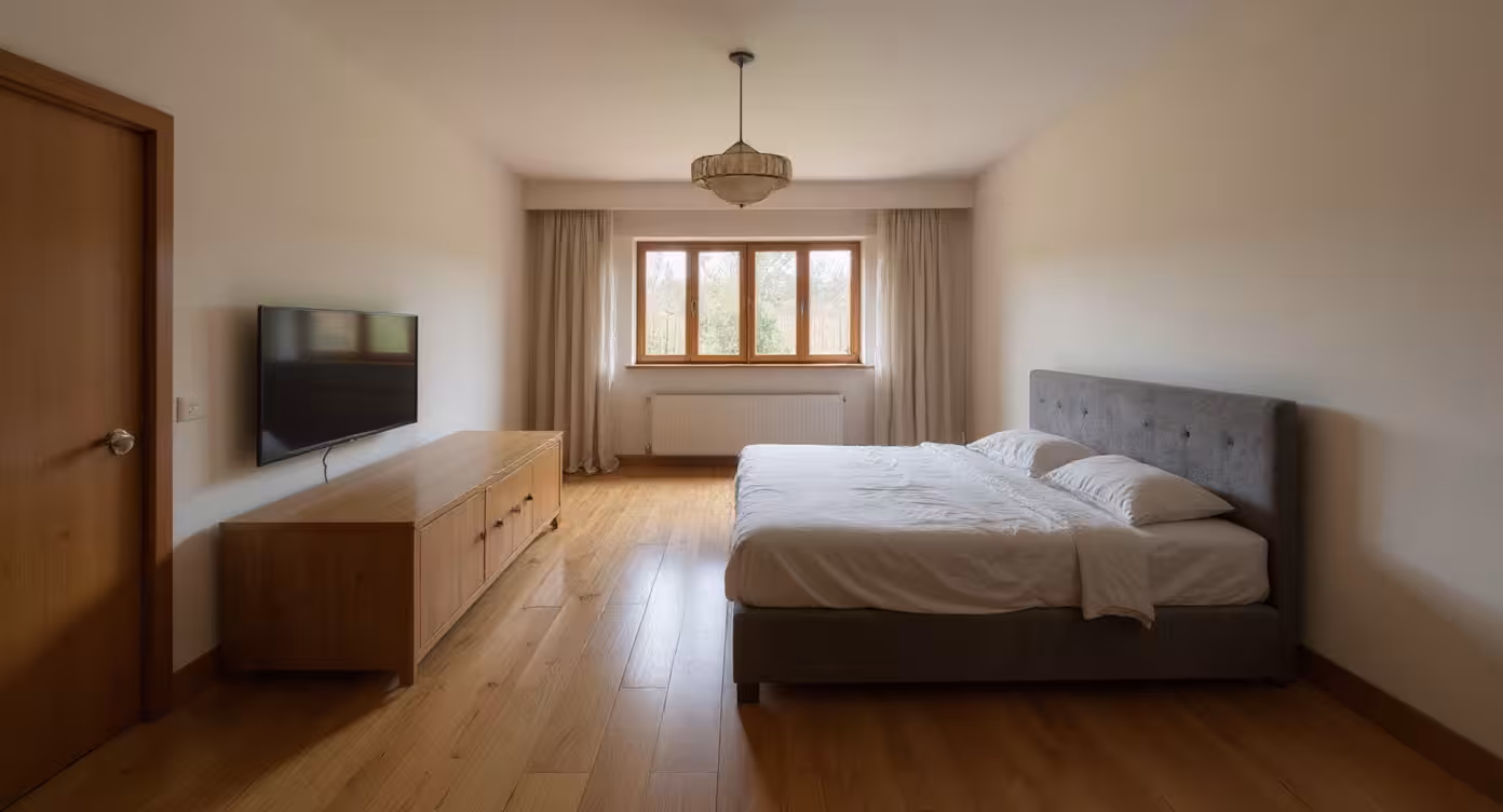 Minimalist bedroom with neutral tones, featuring a gray upholstered bed, wooden flooring, simple wooden cabinet, wall-mounted TV, and natural light from large window.