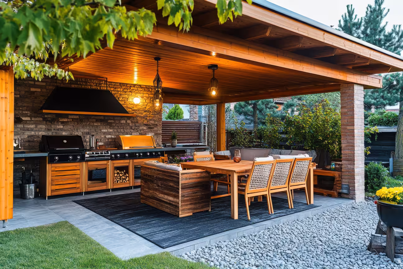 Covered outdoor kitchen dining area with grill station, warm evening lighting, and lush greenery, perfect for relaxed backyard meals and entertaining