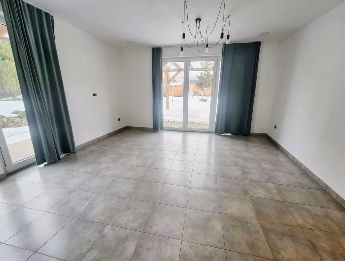 Emptied room with large gray tiled floor, white walls, dark green curtains framing glass doors, and minimalist black pendant light, bright atmosphere.