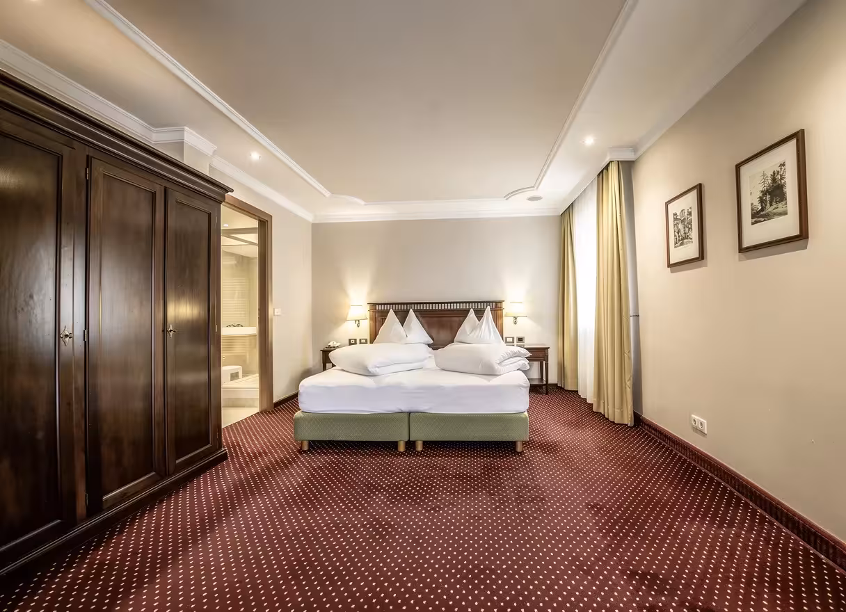 Traditional bedroom featuring a large wooden wardrobe, twin beds with white linens, beige walls, patterned red carpet, framed artwork, and soft warm lighting.