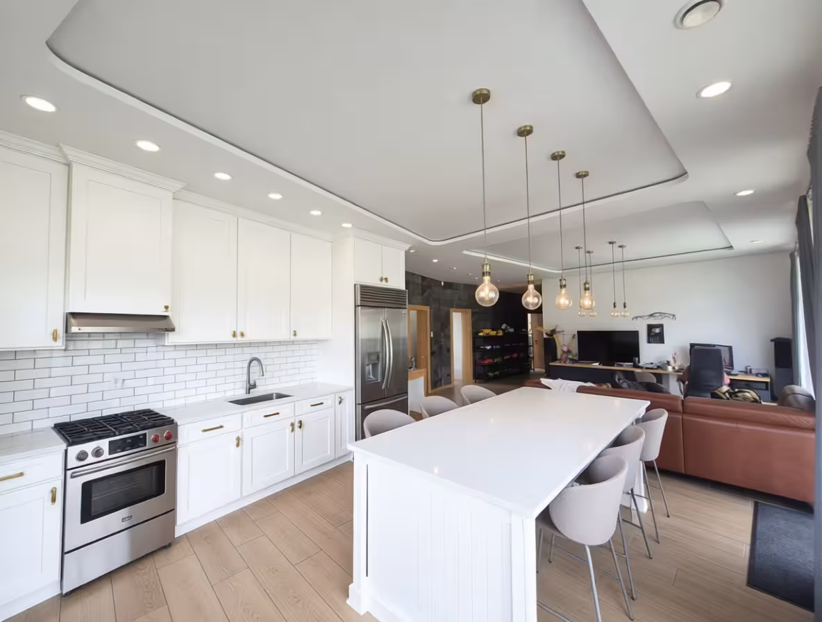 Bright modern open-concept kitchen with white cabinetry, subway tile backsplash, stainless steel appliances, large island with beige chairs, pendant lighting, and open layout to living area.