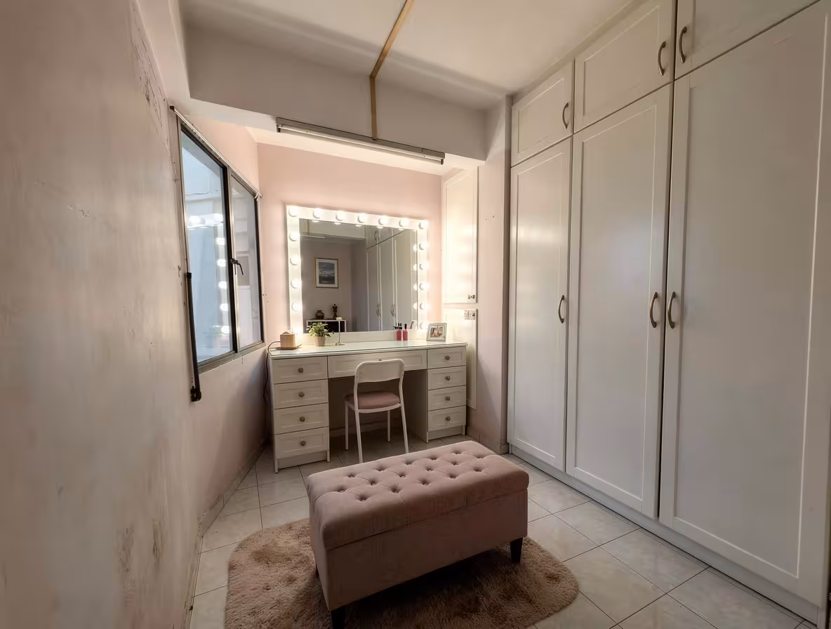 Contemporary dressing room with soft pink walls, white built-in wardrobes, illuminated vanity mirror, tufted ottoman, and neutral rug