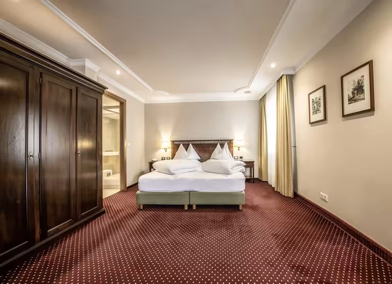 Traditional bedroom featuring a large wooden wardrobe, twin beds with white linens, beige walls, patterned red carpet, framed artwork, and soft warm lighting.