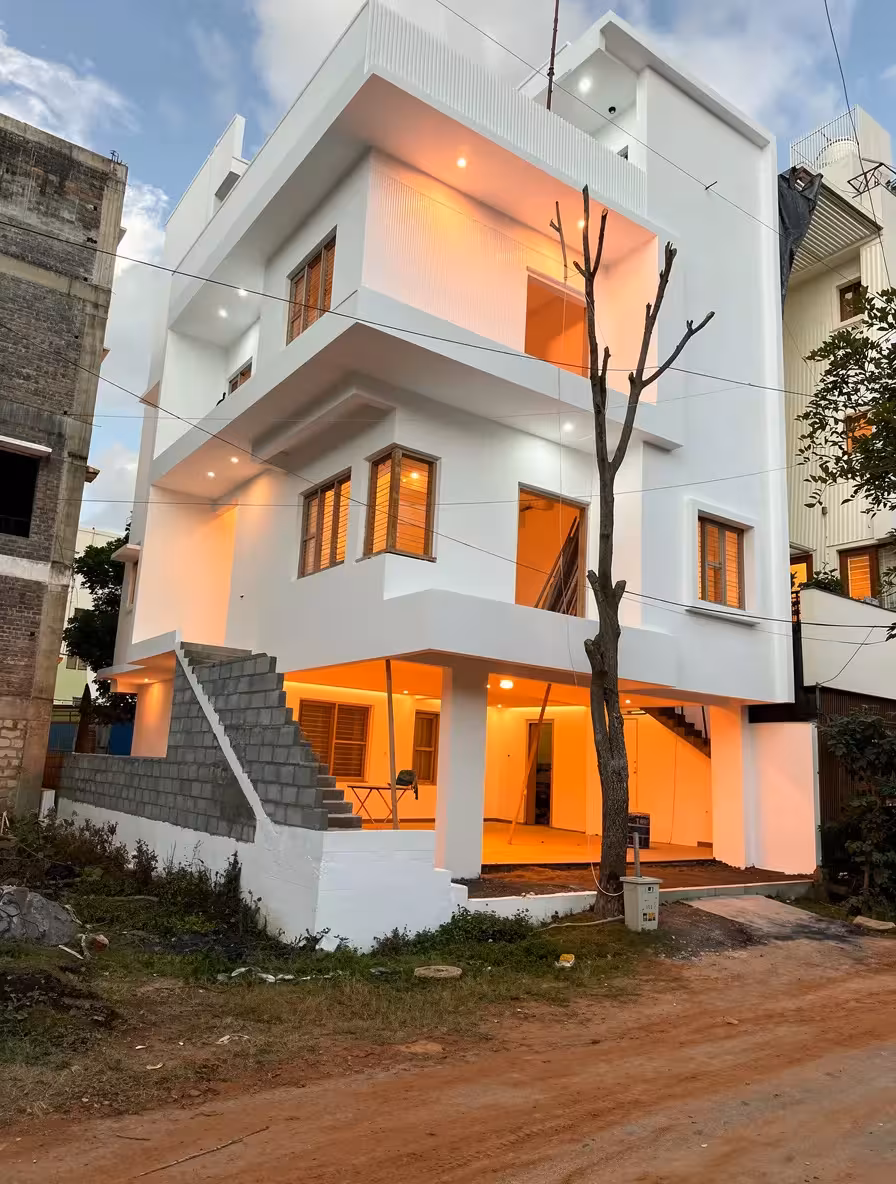 A Contemporary three-story white house with clean lines, warm interior lighting, wooden window frames, outdoor staircase, and a bare tree in the foreground, creating a modern urban atmosphere.