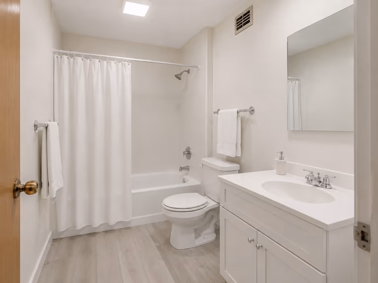 The same dated bathroom after a cosmetic refresh, with painted walls, a white fabric shower curtain, grey vinyl plank flooring and a white shaker vanity
