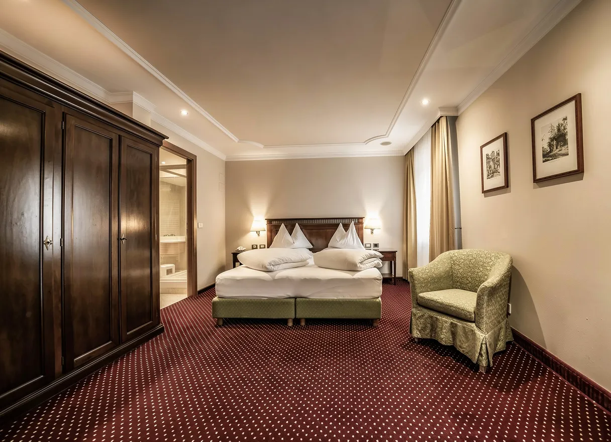 Traditional bedroom with beige walls, dark wood wardrobe and headboard, patterned red carpet, green armchair, soft lighting, and framed artwork.