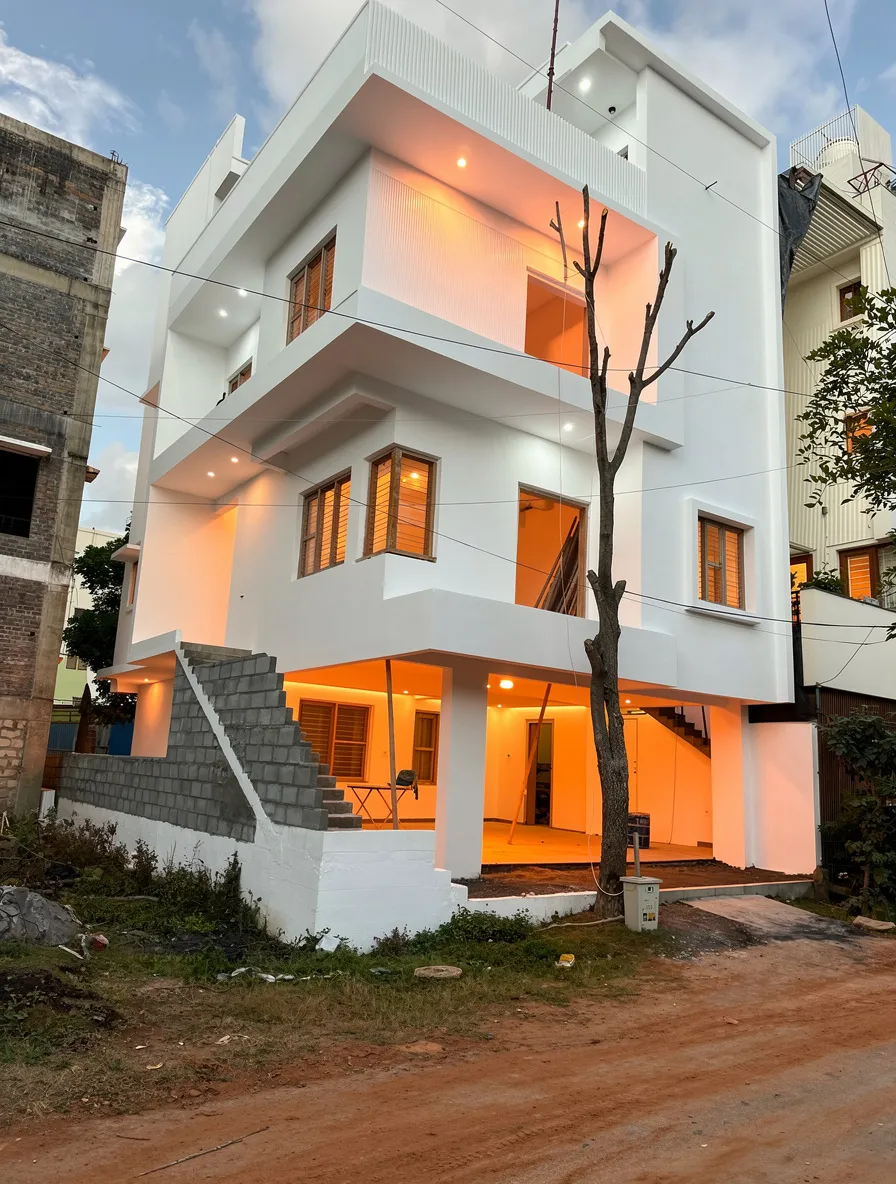 A Contemporary three-story white house with clean lines, warm interior lighting, wooden window frames, outdoor staircase, and a bare tree in the foreground, creating a modern urban atmosphere.