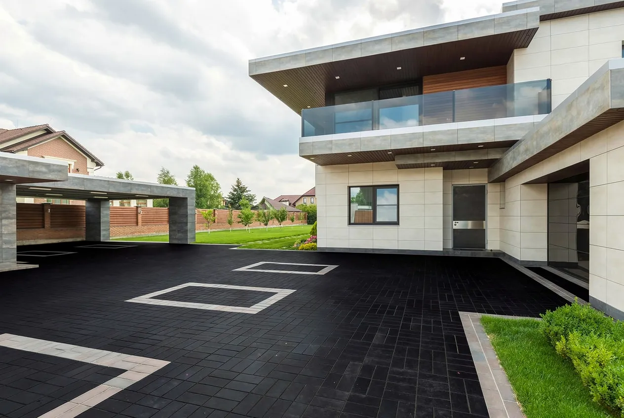 Modern exterior of a two-story house featuring beige stone walls, dark tiled driveway with geometric patterns, glass balcony, and green lawn under cloudy sky.