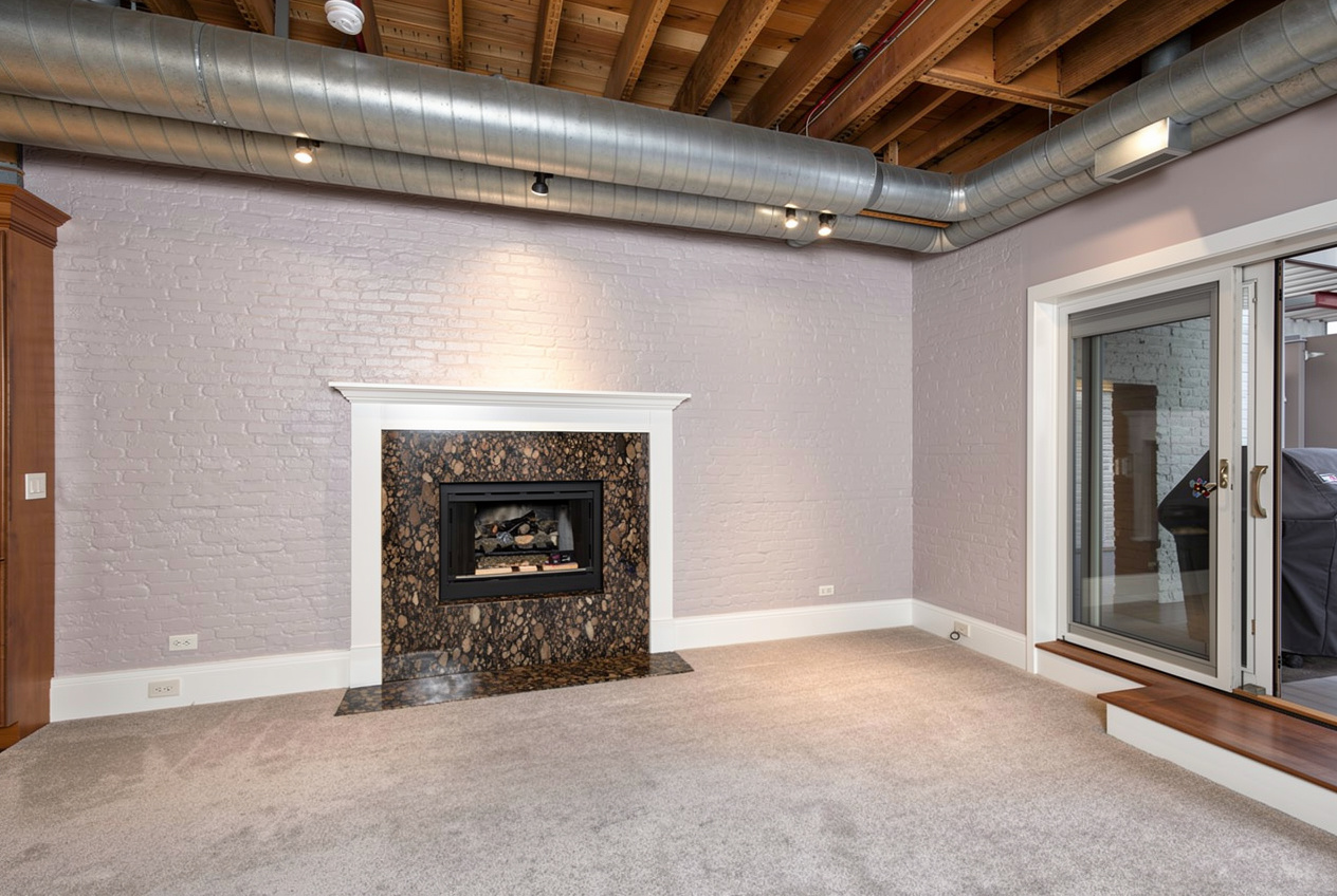 Industrial-style interior room with exposed wood ceiling beams, visible metal air ducts, carpeted flooring, a fireplace with dark stone surround, painted brick walls, and sliding glass doors.