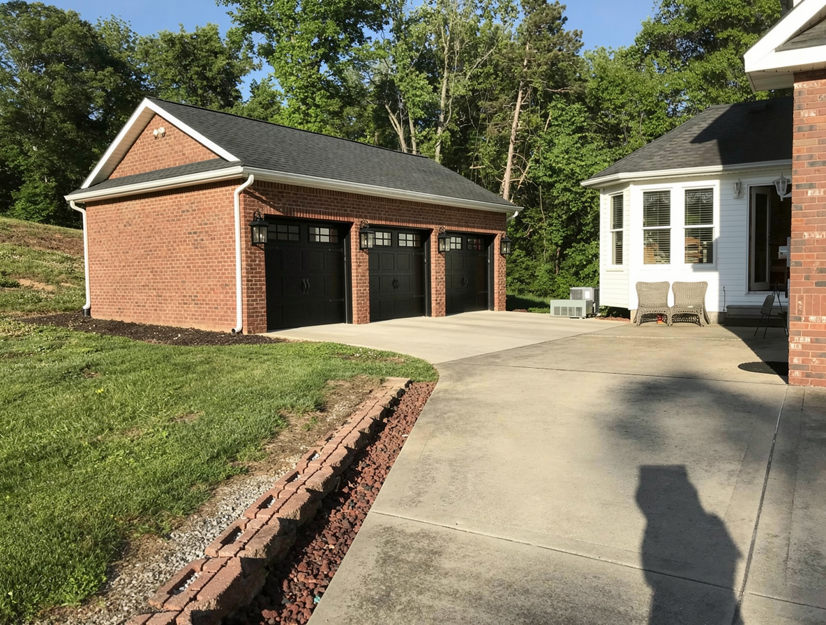 A Traditional exterior design featuring a red brick car garage with black doors, adjacent white house with bay windows, concrete driveway, and lush green surroundings.