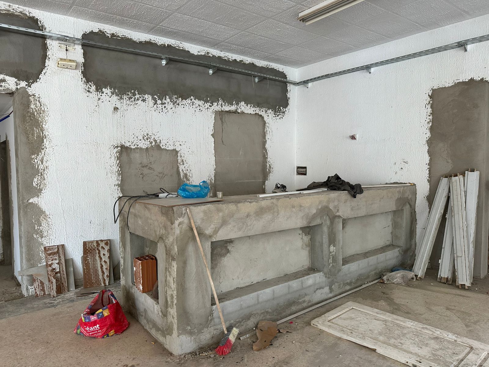 Coffee bar under construction with unfinished concrete counter, rough white and gray textured walls, scattered tools, and raw industrial atmosphere.