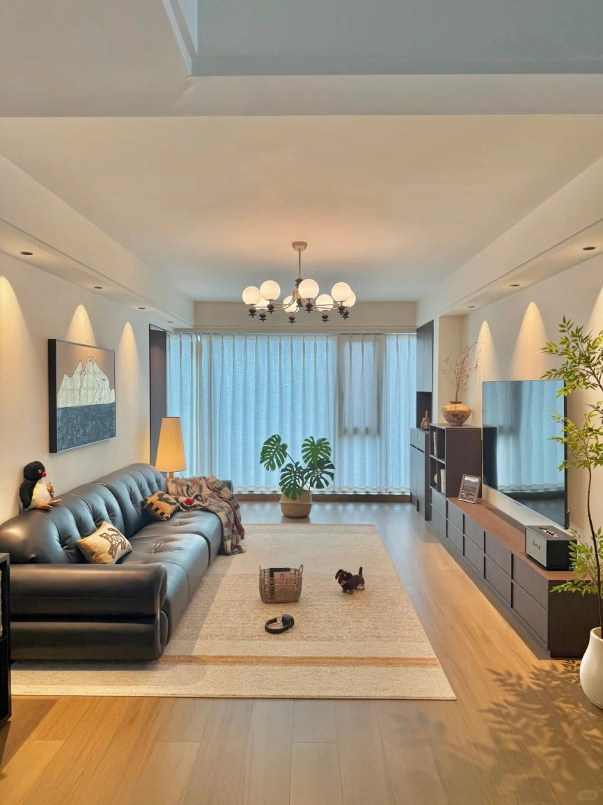 Spacious modern living room with a black leather sofa, beige area rug, wooden flooring, large TV, soft lighting, and neutral tones.