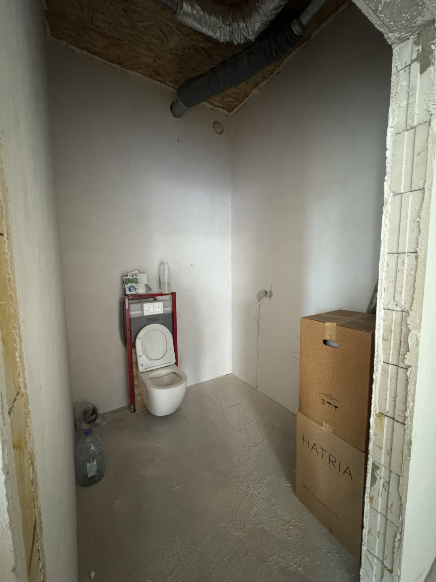 An empty unfinished bathroom with exposed plumbing, bare concrete floor, white unpainted walls, and a wall-mounted toilet frame, conveying a raw, industrial construction phase.