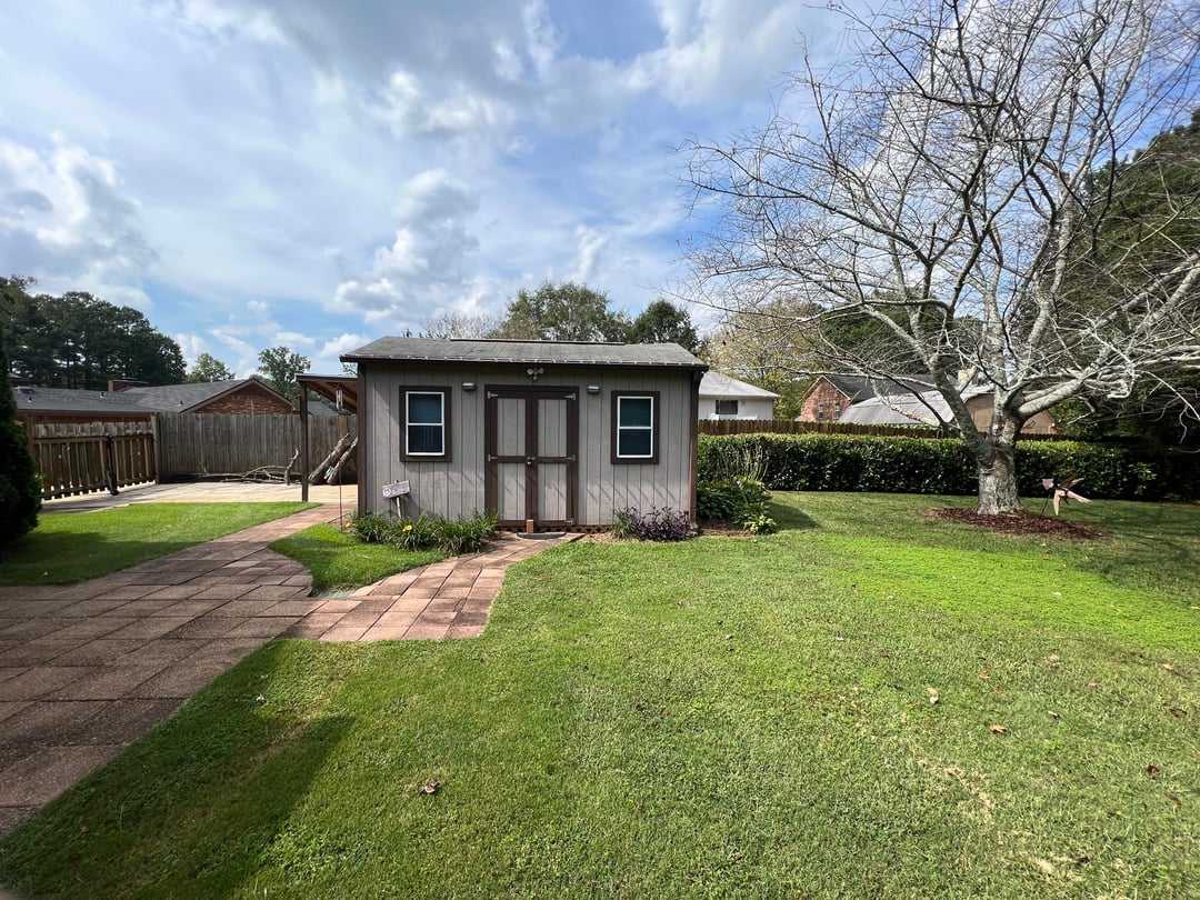 Backyard lawn with storage shed, large tree, brick pathway, and minimal landscaping