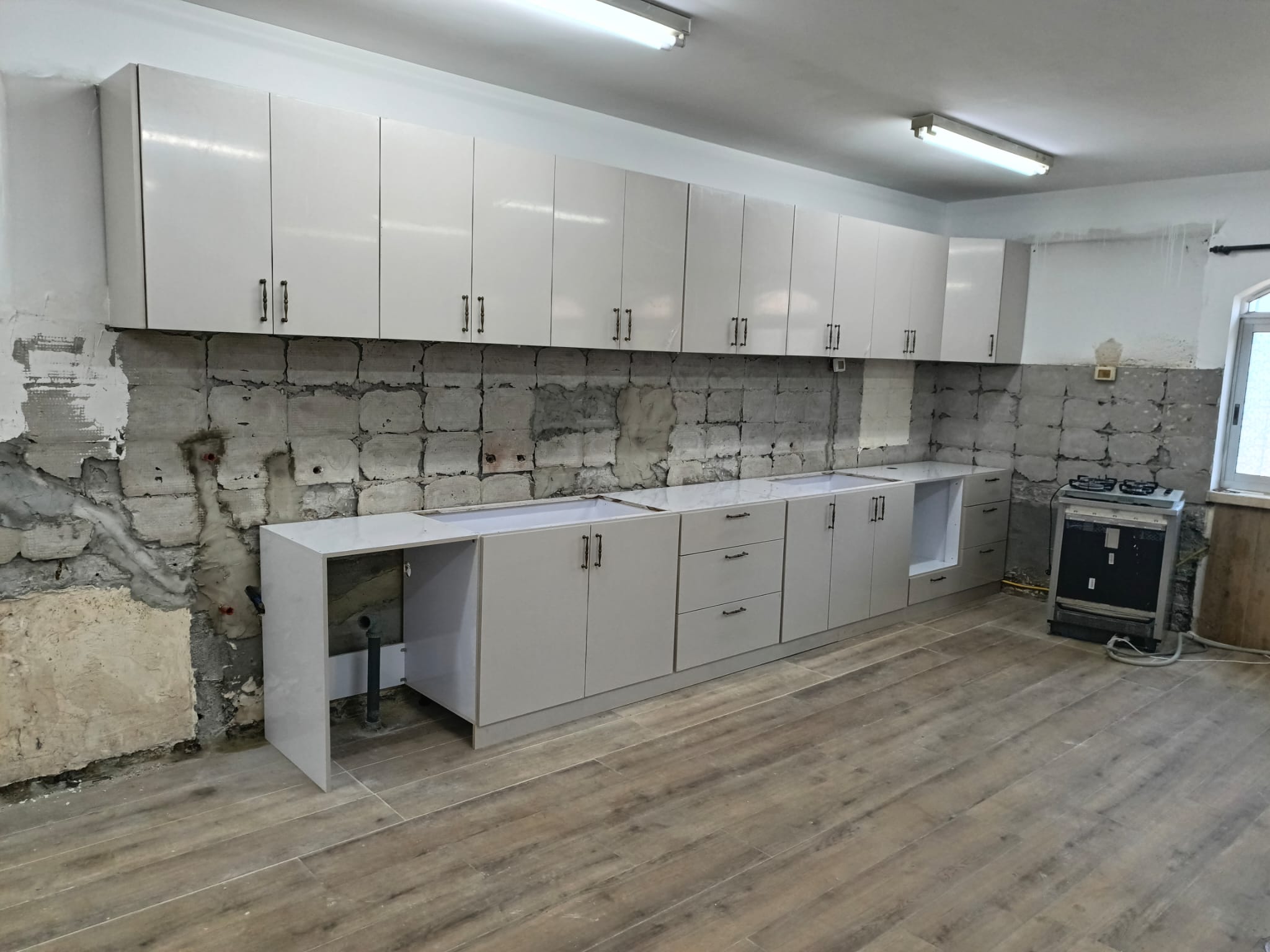 Unfinished single-wall kitchen with white glossy cabinets installed on unfinished concrete walls, wood-look tile flooring, exposed plumbing, and a freestanding gas stove