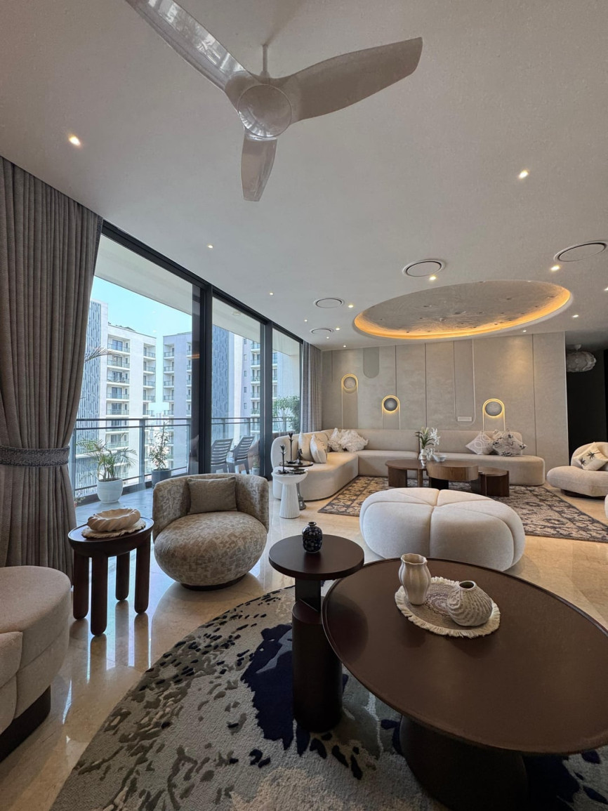 Spacious modern living room with neutral beige and cream tones, curved sofas, textured armchairs, dark wood coffee tables, large windows, and soft ambient lighting.