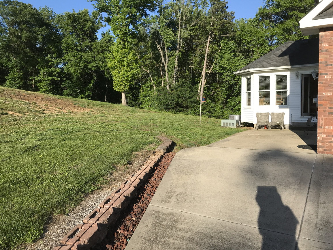 A House exterior area with concrete flooring, two wicker chairs, adjacent to a white and brick house, overlooking a large grassy lawn and dense green trees, peaceful atmosphere.