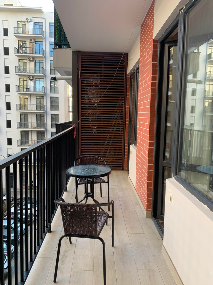 Narrow apartment balcony with black railing, small table set, wood privacy screen, and neighboring buildings