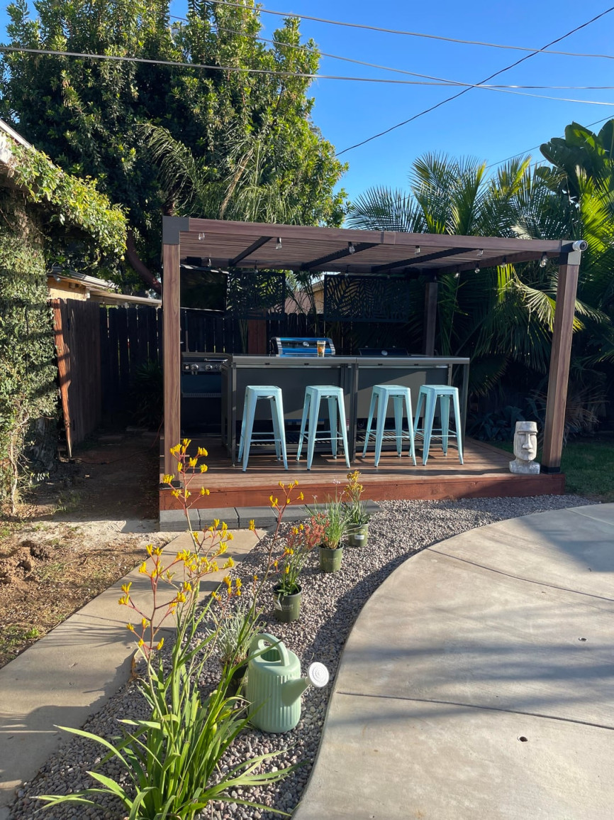 An outdoor backyard bar area with modern pergola, four light blue metal stools, stainless steel counter, surrounded by lush green palm trees and plants, sunny atmosphere.