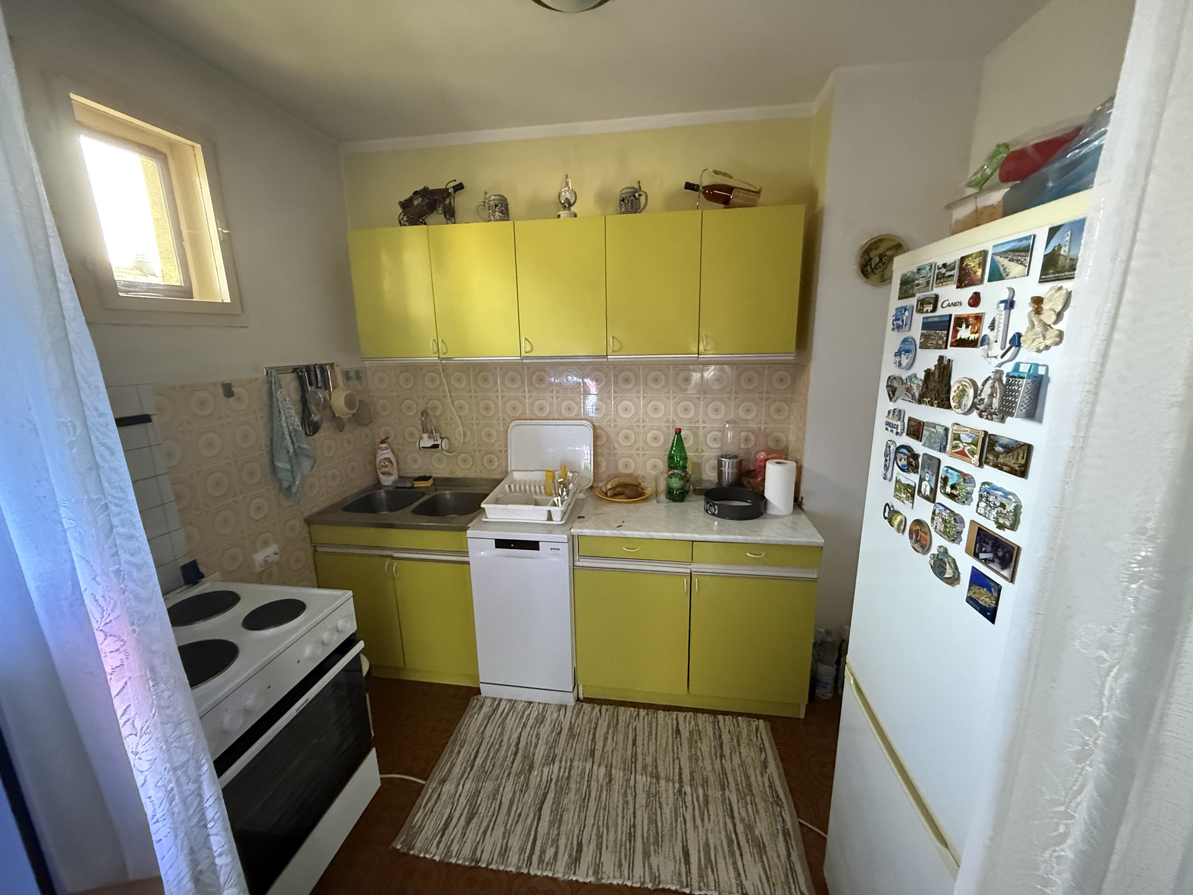 A small vintage U-shaped kitchen with yellow cabinets, patterned beige tile backsplash, white appliances, and a refrigerator covered in colorful magnets, creating a cozy, lived-in atmosphere.