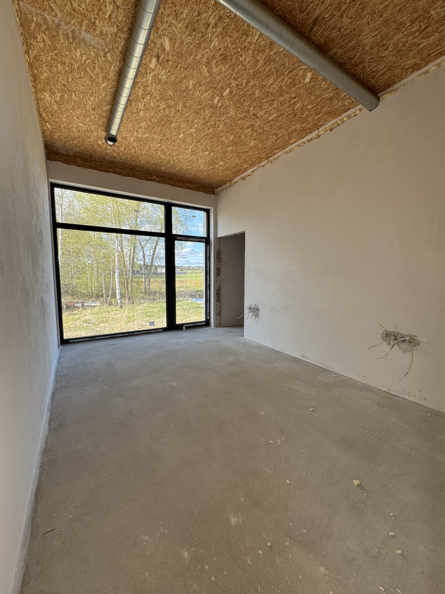 An empty bedroom under construction with raw concrete floor, white plaster walls, large black-framed window, textured wood ceiling, and exposed metal duct, natural light.