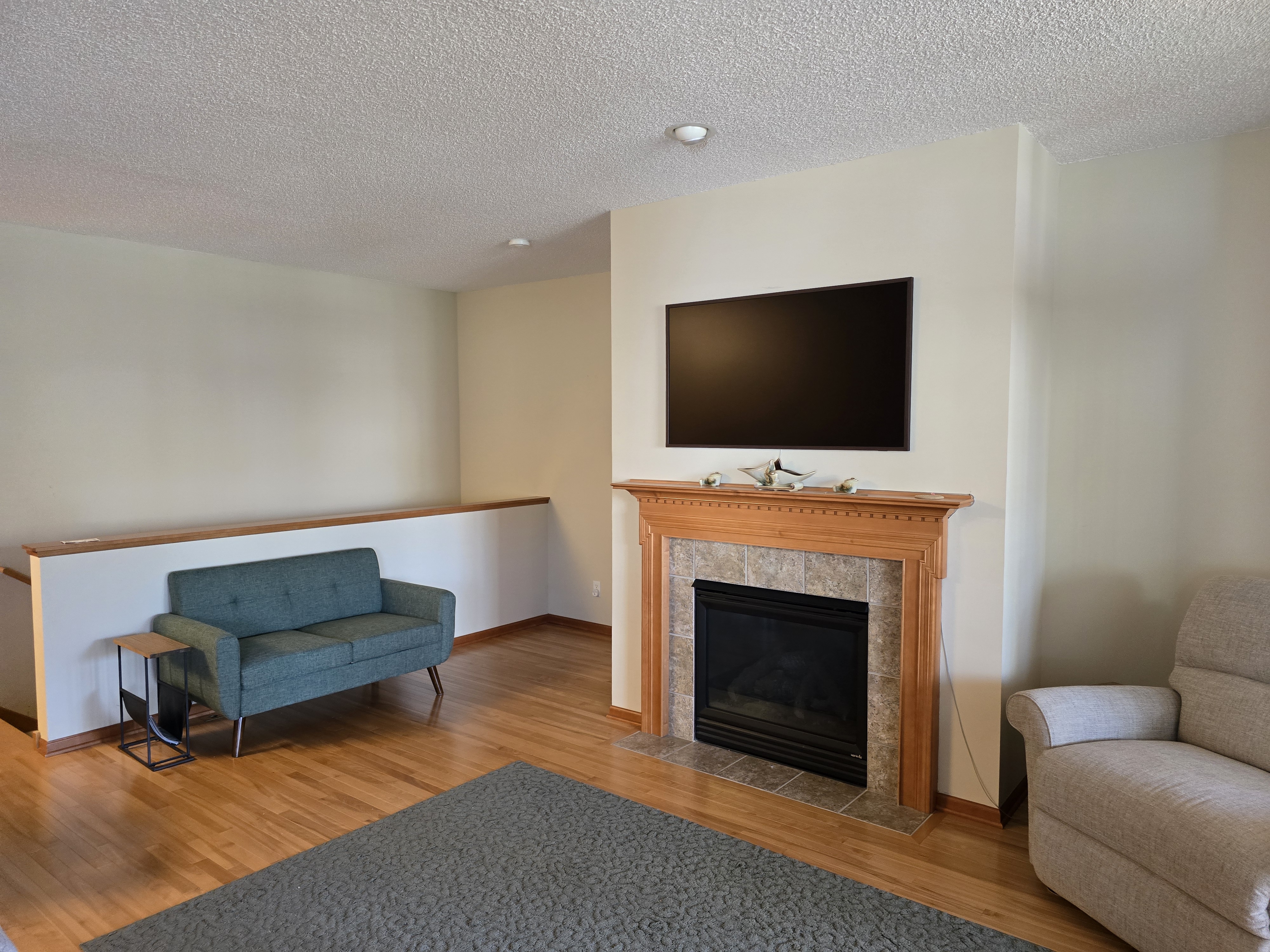 Lliving room with mid-century modern teal loveseat, wooden fireplace with tiled surround, mounted flat-screen TV, beige walls, and warm hardwood flooring.