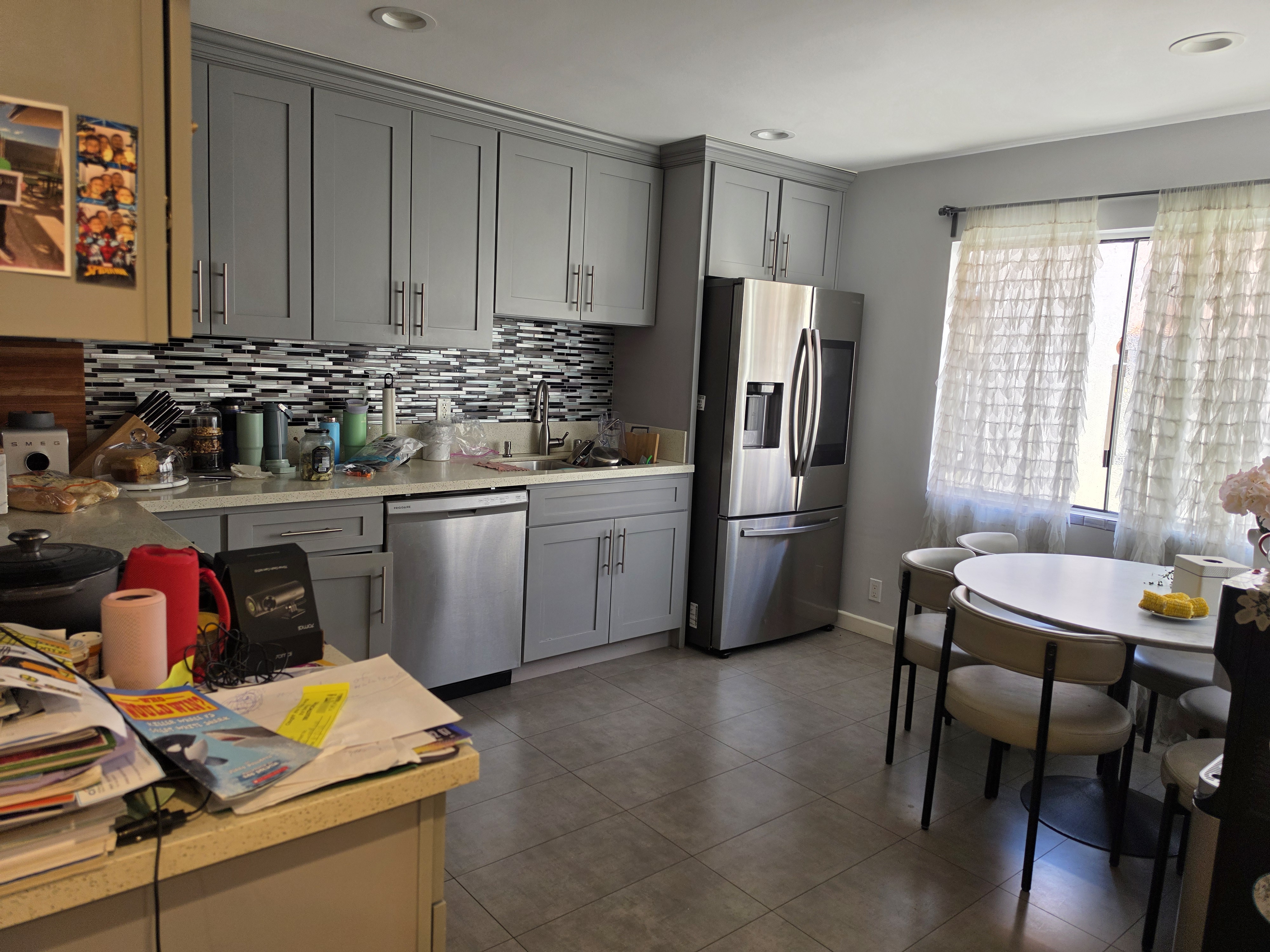 Kitchen with modern gray cabinets, stainless steel appliances, mosaic tile backsplash, cluttered countertops, sheer ruffled curtains, and a casual, lived-in atmosphere.