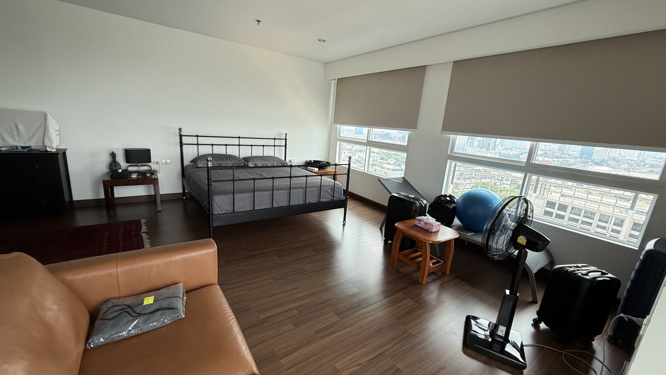 Spacious bedroom with dark wood flooring, black metal bed frame, gray bedding, tan leather sofa, large windows with beige blinds, and scattered personal items.