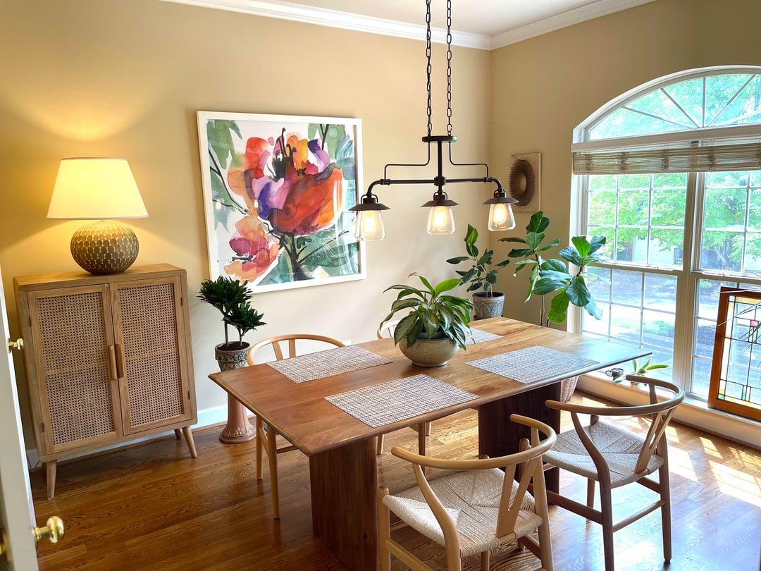 Dining room with wood table, mixed chairs, floral artwork, pendant lighting, and large arched window.