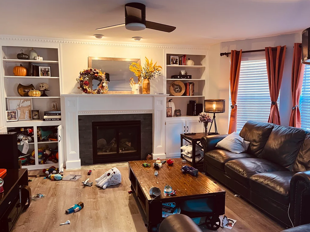 Traditional living room with white built-in shelves, dark leather sofa, wooden coffee table, orange curtains, scattered toys, and warm ambient lighting.