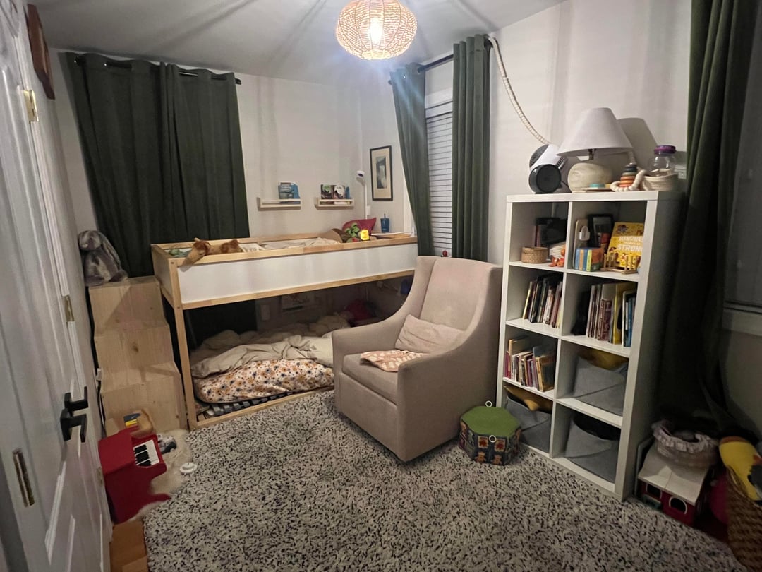 Shared kids bedroom with bunk bed, dark curtains, bookshelf, armchair, and gray carpet