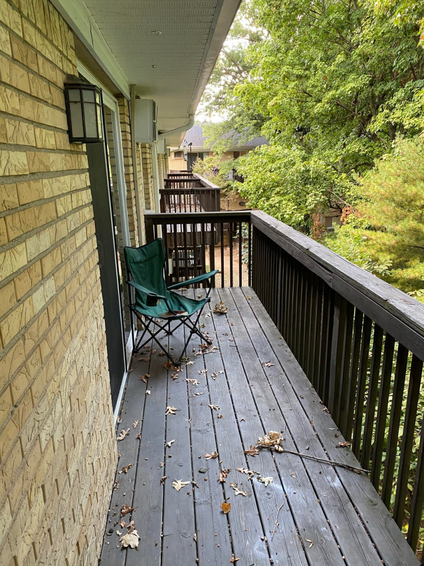 Narrow wooden apartment balcony with folding chair, dark railing, and surrounding trees