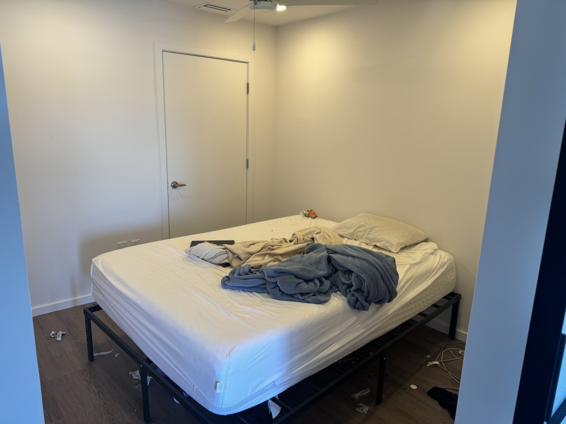 Minimalist bedroom with plain white walls, unmade bed featuring white sheets and blue and beige blankets, simple black metal bed frame, and scattered floor clutter.