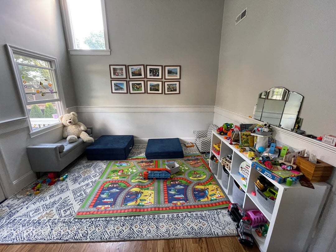 Kids playroom with colorful road rug, toy shelving, blue floor cushions, and gray seating beneath tall windows.