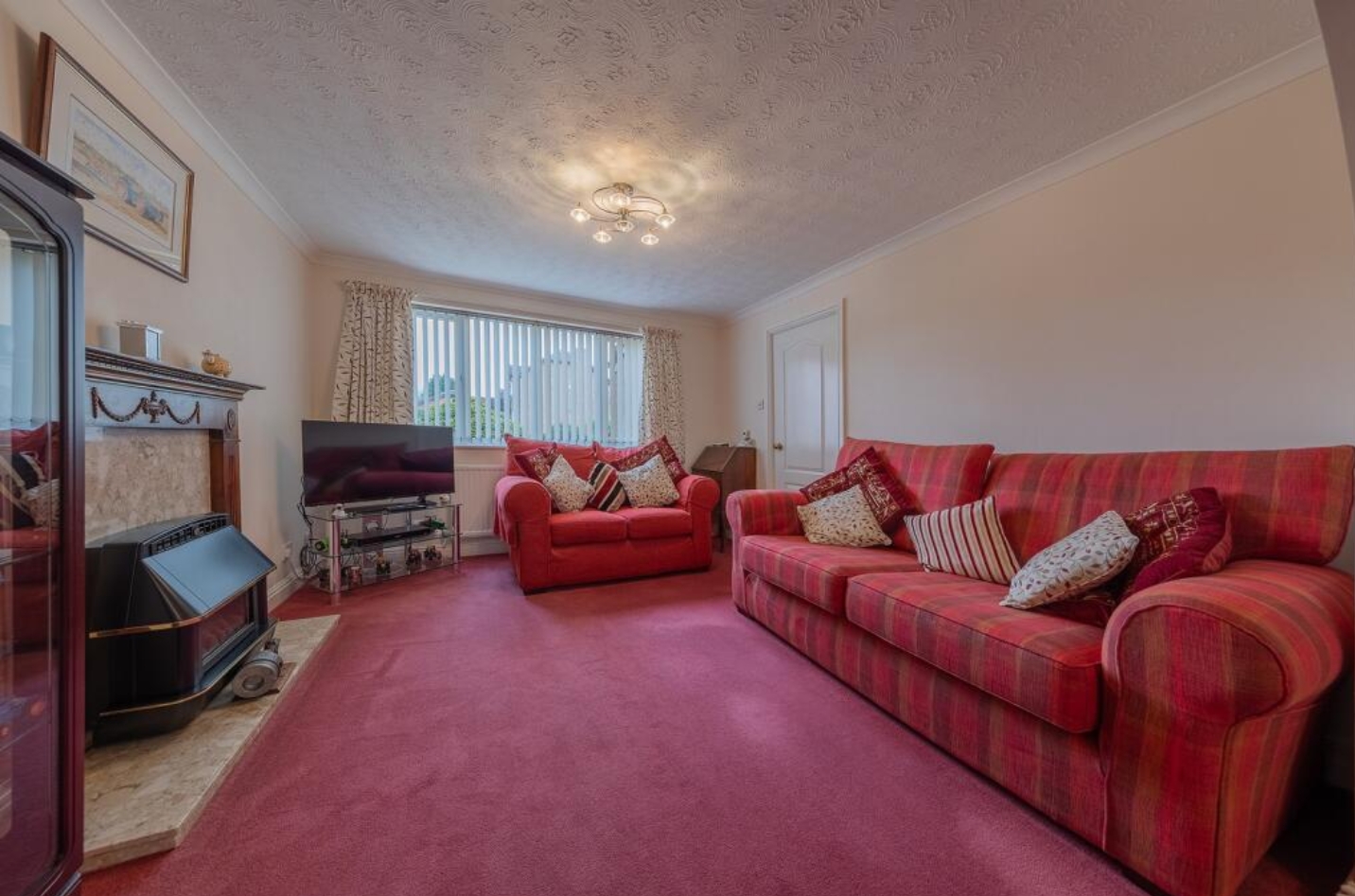 Living room with traditional style featuring red plaid sofas, patterned cushions, a red carpet, beige walls, a fireplace, and a TV under natural light.