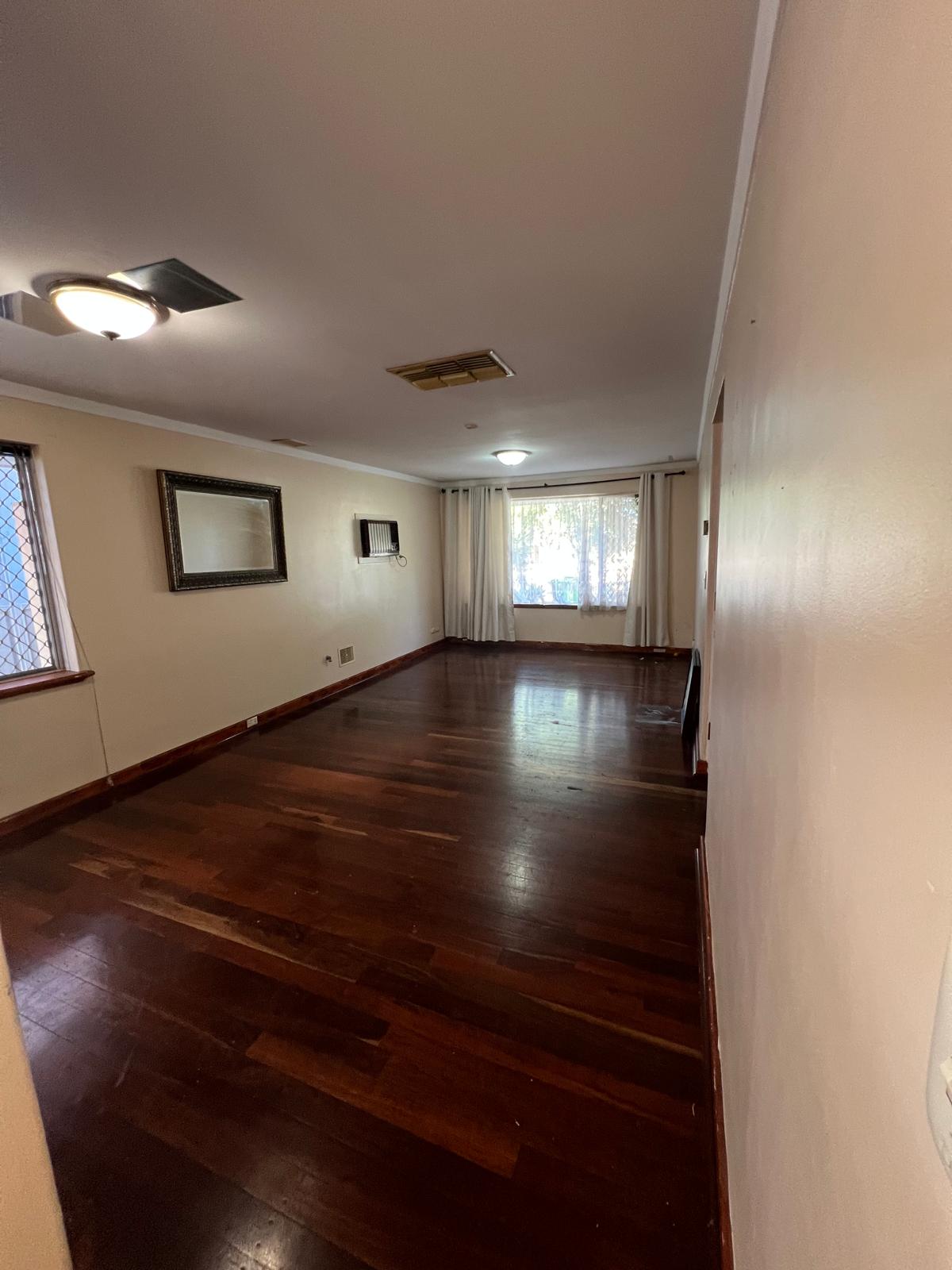 Empty traditional room with dark hardwood floors, beige walls, two ceiling lights, a wall-mounted air conditioner, framed mirror, and large window with white curtains.