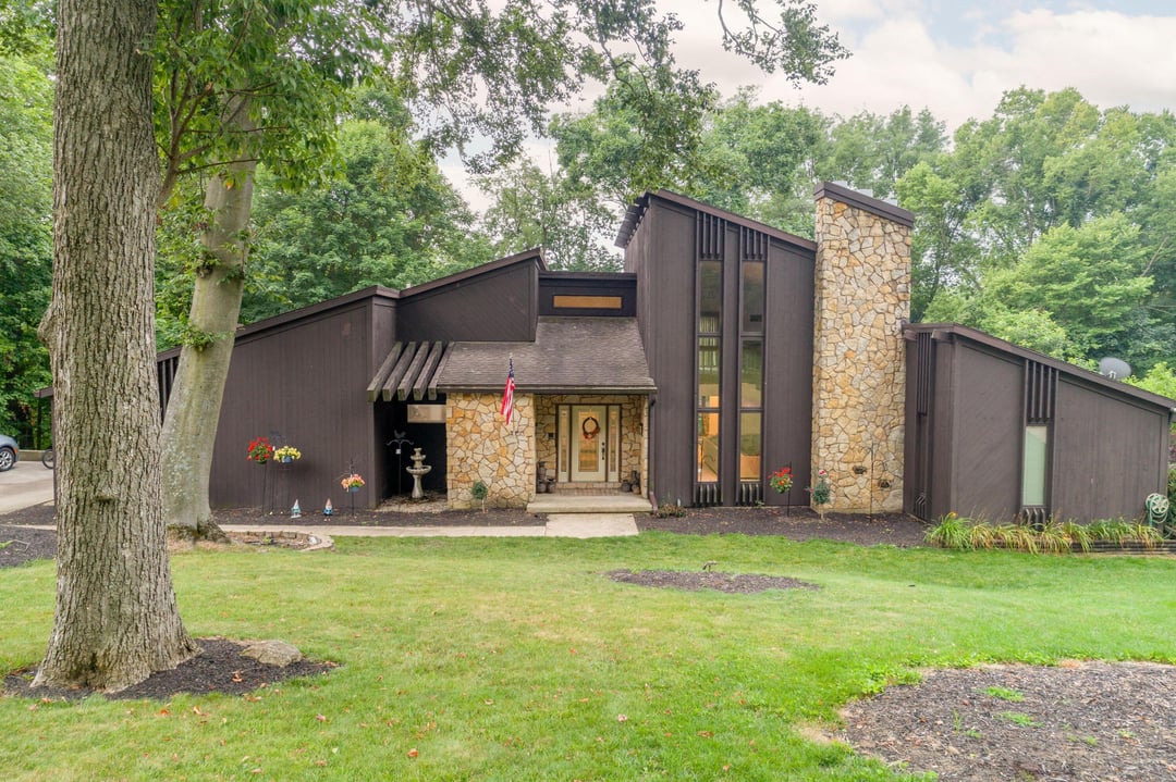 Contemporary woodland home exterior with dark siding, tall stone chimney, and large front lawn surrounded by trees