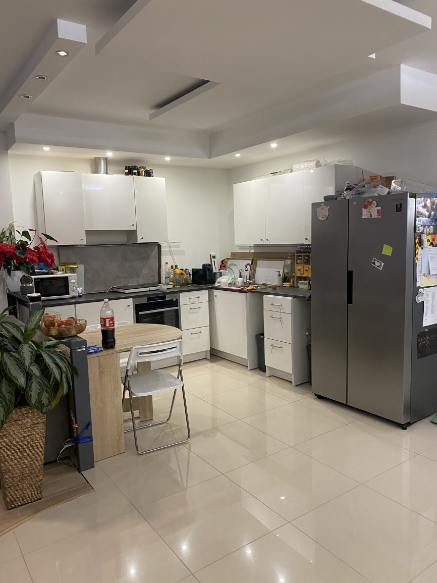 Island kitchen with white cabinetry, stainless steel appliances, beige tiled floor, small wooden dining table, and cluttered countertops