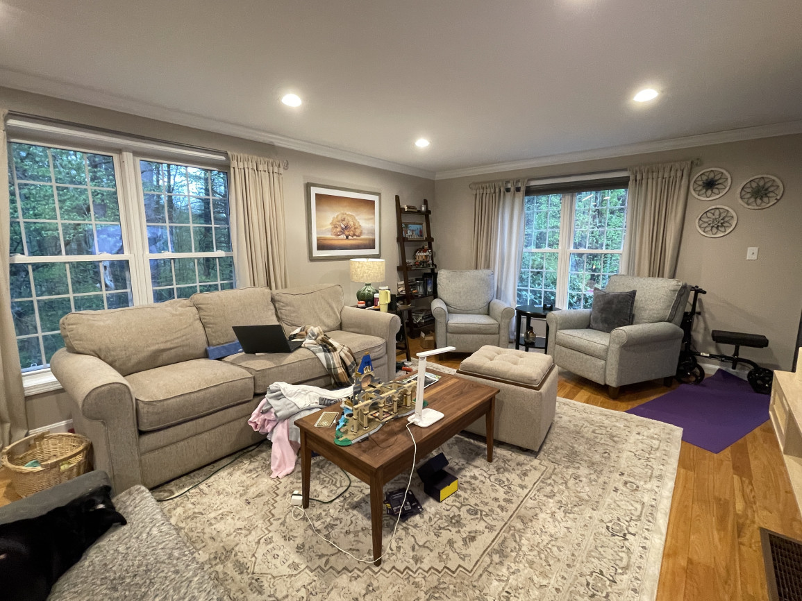 Traditional living room with beige and gray upholstered furniture, large windows with cream curtains, wooden coffee table, patterned rug, and soft warm lighting.